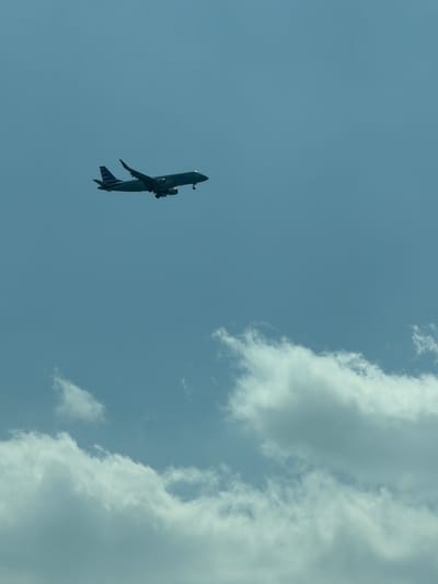 A commercial airplane, identified by the American Eagle logo on its tail, is captured in flight against a clear, pale blue sky. The aircraft appears to be in the process of landing or taking off, with its landing gear visible and extended. Below the airplane, fluffy white clouds are scattered across the lower portion of the frame. The image does not contain any specific landmarks or contextual clues to pinpoint its location within National Harbor, United States of America, nor does it depict any people or interactions.