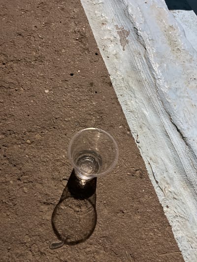 A transparent plastic cup sits on a rough, brownish surface, possibly asphalt or compacted earth, near a white-painted curb or raised edge. The cup contains a small amount of clear liquid, and its shadow is cast on the ground to its left, distorted by the angled sunlight. The surface is textured with small stones and irregularities. A thin white line is visible along the edge of the darker surface, indicating a division or boundary. The scene is outdoors, likely in an urban or semi-urban environment, such as the city of Mashat, Kazakhstan, given the context provided. There are no people or animals present.