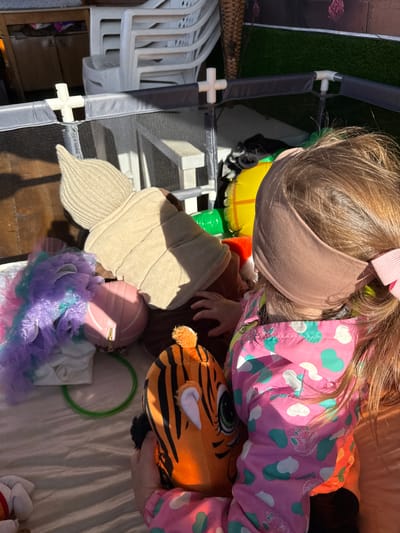 A young child with blonde hair and a pink bow stands indoors, holding a tiger-striped stuffed animal. The child wears a pink jacket with a green and white heart pattern and a brown headband. A tan-colored beanie, a pink purse, and a blue and purple tutu are present on the floor. The indoor location includes a playpen in the top left of the frame, and white plastic chairs are visible nearby. The location is presumably in Кипчак, Russia.