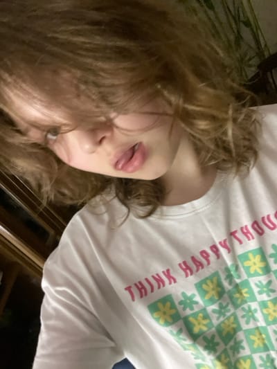 A young person with wavy blonde hair poses in a white shirt with a light green and yellow floral checkered pattern, coupled with the phrase "THINK HAPPY THOUGHTS" in pink lettering. Their tongue is slightly extended, lips are pink, and their gaze is angled to the left of the frame. Background elements suggest an indoor setting with a patterned surface to the upper left and brown wooden panels visible at the bottom.