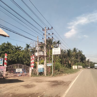 The image captures an outdoor scene in Manetunong, Indonesia, showing a road curving to the right under a clear blue sky with sparse white clouds. The left side of the road features a dirt shoulder, while both sides are lined with abundant green foliage and tropical palm trees.
In the foreground on the left, a metal gate, possibly an entrance to a property, is adorned with numerous red and white flags, characteristic of Indonesia. Partially visible to the left of the gate is a roof structure. A large utility pole stands prominently near the center, equipped with a transformer, insulators, and multiple cross-arms supporting a complex network of power lines that crisscross the upper part of the frame.
Several signs are visible:
- On the utility pole, a white sign reads "YAYASAN PENDIDIKAN BUNDA LINA" (Bunda Lina Education Foundation).
- Below it, a smaller blue sign is present.
- On the gate, a white sign states "KAWASAN BEBAS ASAP ROKOK TK IT BUNDA LINA" (Smoking-free Zone Bunda Lina Kindergarten).
- A partial sign on the far left includes text like "EGRASI NONG ATU RA" and "INDA LINA ID DARU."
The scene is bathed in bright daylight, suggesting a sunny day. In the distance along the road, a white truck and other faint vehicle shapes can be seen. The overall impression is one of a typical rural or semi-urban landscape in a tropical region.
