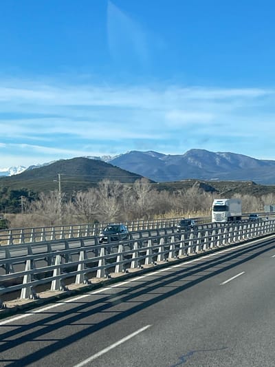 The image presents an exterior scene captured from a moving vehicle on a multi-lane highway, facing northwest toward mountainous terrain near Pont de Molins, Spain. In the immediate foreground, the asphalt road surface shows white lane markings, with long, dark shadows from a barrier stretching across the lanes from left to right, indicating direct sunlight. A sturdy metal bridge barrier, constructed from numerous horizontal and vertical grey-painted beams, runs along the left side of the highway.

On the road beyond this barrier, multiple vehicles are visible in motion. These include a dark blue or grey hatchback, another dark passenger vehicle behind it, and a white semi-truck with a box trailer moving from right to left. Several smaller vehicles are visible in the far distance on the same road.

The midground landscape behind the bridge barrier features a flat area covered with numerous bare, leafless trees and shrubs, suggesting a winter season or arid conditions. A brown utility pole with overhead power lines stands amidst this vegetation. The background is dominated by a range of dark mountains, some of which exhibit patches of snow on their higher peaks, particularly towards the left horizon. The sky is bright blue with scattered, thin white cloud formations, predominantly in the upper half of the image. No individuals are discernible in the scene.