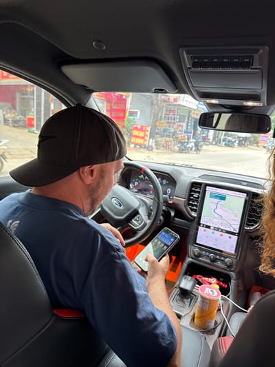 The image captures the interior of a car parked on a street in Bắc Yên, Vietnam. A man wearing a dark baseball cap and a blue t-shirt is seated in the driver's seat, holding a smartphone in his left hand. The car's dashboard features a large touchscreen display showing a navigation map, and a digital instrument cluster. A cylindrical container of snacks is visible in the center console. The street outside appears to be busy, with buildings lining the road and several motorcycles parked. The overall scene suggests a moment of pause during a journey, with the driver possibly checking his phone or the navigation system.