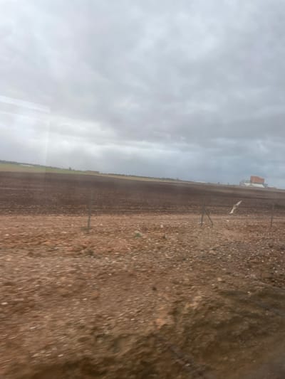 A tilted view of a field under a cloudy sky. The field is mostly brown with some green patches along the edge. A few wooden posts and a white marker stand in the field. A building can be seen in the distance. The overall impression is of a rural scene on a somewhat overcast day.