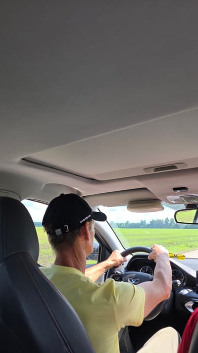 An outdoor, daytime shot captured from inside a vehicle, looking forward. The scene focuses on a person driving, viewed from the backseat of the vehicle. The driver, wearing a black baseball cap and a light yellow t-shirt, is positioned at the steering wheel. Their hands are on the wheel, and their arms are visible. The interior of the car includes a black dashboard and steering wheel with a visible emblem, as well as a sunroof and a rearview mirror. Outside the car, a bright green field stretches to the horizon, lined with trees. The sky is light and appears to be clear. The image does not contain enough information to determine a specific location within Dar'ino, Russia.