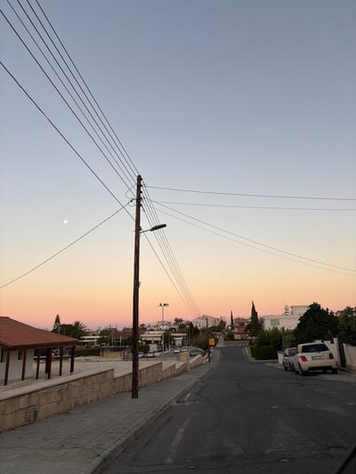 This is a medium-shot, vertical, eye-level, outdoor photograph of a street in Kolossi, Cyprus, at dusk. The sky is a gradient of pale blue at the top fading into soft pink and orange hues towards the horizon. A full moon is visible in the upper left quadrant of the sky.
Dominating the foreground and midground on the left side of the image is a paved sidewalk and a low stone wall. To the left of the sidewalk is a building with a reddish-brown tiled roof and white walls, supported by wooden pillars. The street, paved with asphalt, curves gently uphill towards the right side of the frame.
A tall, dark wooden utility pole stands prominently in the center of the image, supporting multiple overhead wires and a streetlamp. Several electrical wires crisscross the frame against the sky.
On the right side of the street, several vehicles are parked. The closest vehicle is a light-colored hatchback with license plate KRL-934. Behind it, another car is partially visible. Houses and buildings with white walls and various roof types line the street. Some trees, including a tall, slender cypress, are scattered among the buildings. A speed limit sign indicating "50" is visible on the left side of the road in the distance. The overall scene depicts a quiet residential street at sunset.