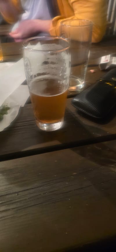 A half-empty glass of amber-colored beer with foam sits on a dark wooden table next to an empty glass and a black wallet or pouch. There is white napkin on the table as well. Part of a yellow shirt and blue shirt is visible along the edge of the table. The location appears to be a pub or restaurant, based on the beer and setting, but there is no information on the city.