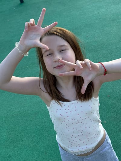 A young girl with brown hair and closed eyes, wearing a white tank top with pink hearts and grey shorts, stands on a green surface, possibly a playground. Her hands are held up, framing her face in a gesture that resembles a camera or a peace sign. She is wearing a clear, beaded bracelet on her left wrist and a red string bracelet on her right wrist. The background is a solid green texture, suggesting an outdoor setting. The image appears to be a candid shot, taken from a slightly elevated perspective. The location is Boksitogorsk, Russia.
