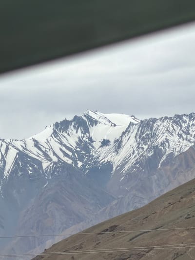 The image shows a mountainous landscape in Pidmo, India, under a cloudy sky. In the foreground, a dark, angular object partially obstructs the view from the top, casting a shadow over the scene. Below this, a sloping, barren hillside dominates the lower half of the frame, with two thin, parallel lines suggesting overhead wires. The middle ground is occupied by a range of majestic, snow-capped mountains, their jagged peaks and steep slopes etched with shadows and streaks of snow. The overall impression is one of rugged natural beauty and vastness.