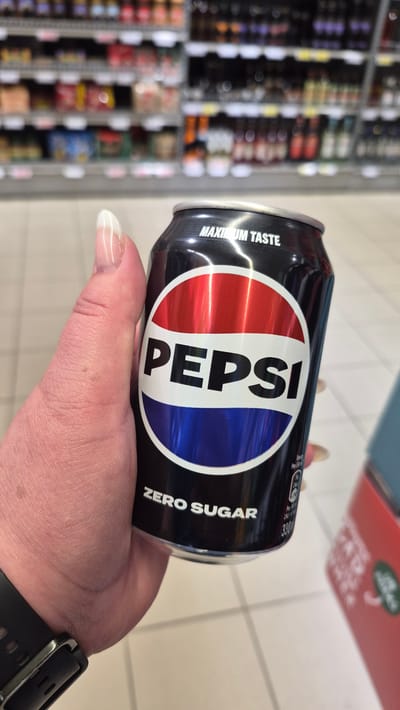 A person's hand holds a can of Pepsi Zero Sugar. The can is black with the iconic Pepsi logo, which is a red, white, and blue circle. Text on the can reads "MAXIMUM TASTE" and "ZERO SUGAR". The background appears to be a grocery store aisle with shelves stocked with various products. The floor is tiled. The scene is indoors.