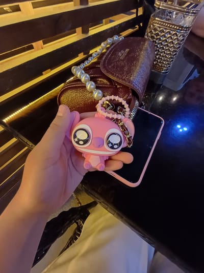 The image shows a close-up of a person's hand holding a pink, cartoonish keychain. The keychain has large, wide eyes with black pupils and white highlights, giving it a startled or surprised expression. It also features small, rounded pink ears and what appear to be stubby pink limbs. A gold chain connects the keychain to a loop, suggesting it's designed to be attached to a bag or keys.
In the background, a shiny, dark brown purse with a textured surface is visible. A string of faux pearls is draped over the purse. Next to the purse is a pink smartphone case, with the phone itself obscured. A decorative, metallic object with an intricate, geometric pattern is also present on the left side of the image. The overall setting appears to be indoors, possibly at night or in a dimly lit area, with some yellow and blue lights casting reflections on the dark surfaces. The surface the objects are resting on appears to be made of dark wood with thin yellow stripes.
The scene is focused on the hand and the keychain, suggesting the keychain might be a new purchase or a favored accessory. The lighting and the presence of a purse and phone imply a casual or social setting. No specific activity or event is clearly depicted, but the emphasis on personal items could suggest someone is preparing to leave, or simply showcasing their belongings. The location context of Nematjan, Uzbekistan, is not visually evident in the image itself.