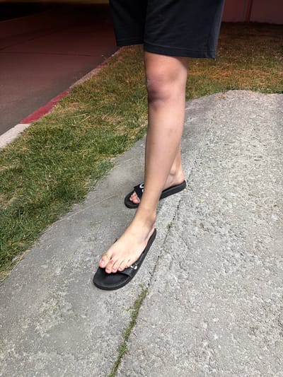 A person's legs, wearing black shorts and black slides, stand on a concrete sidewalk next to a patch of grass. The sidewalk has visible cracks and texture. In the background, a road with a red curb is visible, and the lighting suggests it is nighttime.