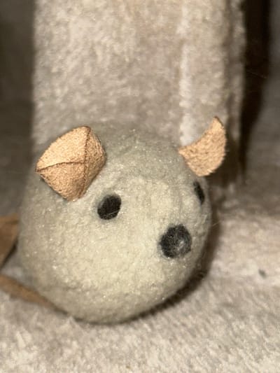 The image is a close-up, slightly blurry photograph of a small, stuffed toy mouse. The mouse is light gray and has two small, tan, triangular ears, one on each side of its head. It has two black bead-like eyes and a small, black, stitched nose. The mouse is sitting on a textured, light-colored surface, which appears to be fabric, possibly a carpet or upholstery. Behind the mouse, a blurred, vertical, light-colored object is visible. The lighting in the image is soft, creating some shadows. The location in the city of Edmonton, Canada, cannot be determined from the visual information provided in the image.