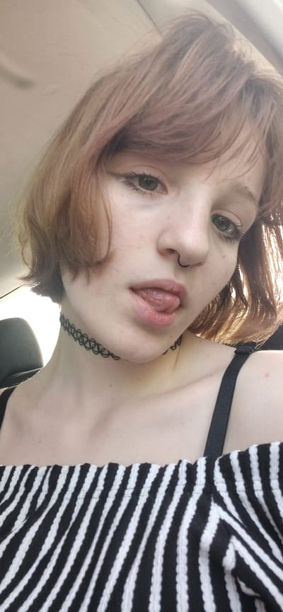 A young woman with light skin and auburn hair is seen in a close-up shot. She is wearing a black choker necklace and a black and white striped top. She has a nose ring and her tongue is slightly protruding. The background appears to be the interior of a car. The location of the scene is not visibly discernible from the image and cannot be confirmed to be Bistrice, Montenegro.