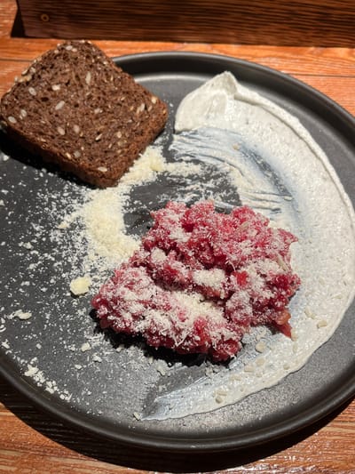 A close-up, high-angle shot depicts a prepared dish served on a dark, matte ceramic plate resting on a wooden table surface. The dish consists of a mound of finely minced, bright red raw meat, possibly beef tartare, positioned towards the center-right of the plate. This meat is heavily garnished with a fine layer of grated white cheese. To the upper left, a single, rectangular slice of dark brown, seedy bread, likely rye or whole grain, is present. A prominent, curved swath of thick, off-white creamy sauce with some visible texture is spread along the right side of the plate, partially underlying the meat. Additional grated white cheese is scattered across the dark plate surface between the bread and the meat. The wooden table beneath exhibits a visible grain pattern. No individuals are present in the image. The specific location within Родниковское сельское поселение, Ukraine, cannot be determined from the visual information provided.