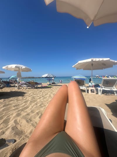A first-person perspective shot captures a sunny day at a crowded beach in Protaras, Cyprus. The foreground shows a person's tanned legs and green bikini bottoms resting on a beach chair, with sand visible in the lower left corner. Behind them, numerous beach umbrellas dot the sandy shore, providing shade for beachgoers. Several lounge chairs are occupied by people sunbathing or relaxing. The sea, a clear blue, stretches out to the horizon, with many people swimming and enjoying water activities. Jet skis and other watercraft are visible further out. The sky is a cloudless, vibrant blue. The overall scene conveys a typical summer day at a popular tourist beach.