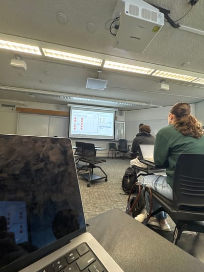The image displays an interior view of a classroom or lecture hall located in Wellesley, United States. From a student's perspective, a silver laptop (MacBook Pro) with a dark, reflective screen occupies the lower left and center foreground, partially obscuring the view of the room. Its keyboard is visible. In the middle ground, three individuals are seated at individual desks with integrated chairs, facing a white projection screen at the front. The closest visible student, positioned on the right, wears a green long-sleeved shirt and light blue jeans, with reddish-brown hair tied back. An open laptop is on their desk. To their left, another student, wearing a dark hoodie, also has an open laptop on their desk. A third student is partially visible further in the background. A white Panasonic projector is mounted on the light-colored acoustic tile ceiling, actively projecting a calendar displaying "October 2025" and "November 2025" with highlighted dates onto the screen. The ceiling features multiple long fluorescent light fixtures. The floor is covered with a patterned gray carpet. A black backpack and a dark tote bag are situated on the floor near the seated students.