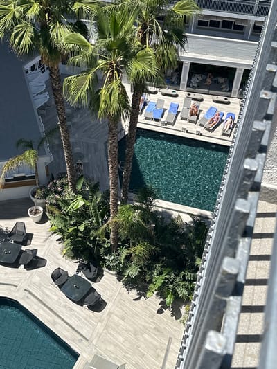 This is an overhead shot of a resort area in Lloret de Mar, Spain. The scene depicts a swimming pool and a poolside lounging area. Several people are sunbathing on lounge chairs arranged on a wooden deck. Some are covered with blue towels. Palm trees and lush greenery are scattered throughout the area, casting shadows on the tiled ground. Outdoor furniture, including tables and chairs, is set up on the deck, suggesting an area for dining or relaxation. The water in the pool is dark and appears to be rippling. The lighting suggests it is a sunny day.