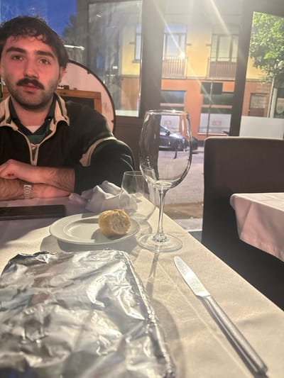 A man is sitting at a table in a restaurant located at 50.854, 4.386 in Schaerbeek, Belgium. He is wearing a black jacket with a white stripe. His arms are crossed, and he is looking directly at the camera. There is a wine glass and a half-eaten piece of bread on the table. A knife is also on the table, lying next to a white napkin. There is a large piece of aluminum foil on the table, and a small glass in the background. The table is white, and there is another table next to it, also covered in a white tablecloth. The restaurant has large windows that look out onto a street. There is a brown chair to the right of the table. A car is visible through the window.