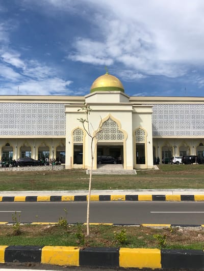 The image captures a contemporary mosque located in Reuleut Timu, Indonesia. The mosque's facade is a light yellow, accented by ornate white latticework above a series of arched entrances. A prominent golden dome with a green base crowns the central section of the building, topped by a crescent moon and star. Several vehicles, including black SUVs and a white SUV, are parked in front of the mosque, with a group of people, some wearing green, gathered near the entrance. The mosque is situated behind a lawn and a low stone wall, and in the foreground, a road is visible with distinctive yellow and black striped curbs. The sky above is a vibrant blue, dotted with white, scattered clouds. A slender, leafless tree stands in front of the mosque, positioned between the building and the road.