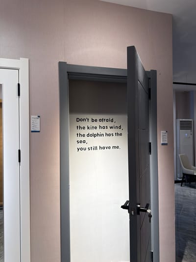 The image shows a partially open door with a message on it. The door is dark gray and has a doorknob with a key in it. The message on the door reads, "Don't be afraid, the kite has wind, the dolphin has the sea, you still have me." The door is set into a wall that has a beige, textured surface. Below the dark gray door is a white door. There are two small information labels on the wall, one above the gray door and one between the two doors. The door appears to be in a showroom or exhibition space, as the background includes glimpses of other exhibits, including an air conditioner unit and a white chair. The location is likely in Chancheng District, China, based on the text "gorge" being recognized which is one of the texts from the information labels in the image.