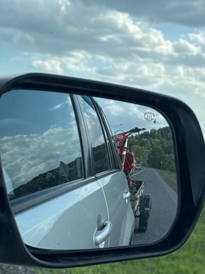 The image shows a side mirror of a vehicle reflecting a red dirt bike on a trailer being towed. The trailer has a black box-like object on it. The road is asphalt, with greenery visible in the background. The sky is visible as well with white puffy clouds. The vehicle door is also visible within the mirror's view. There is no evidence to suggest the image's location is in Ir'yak, Russia.