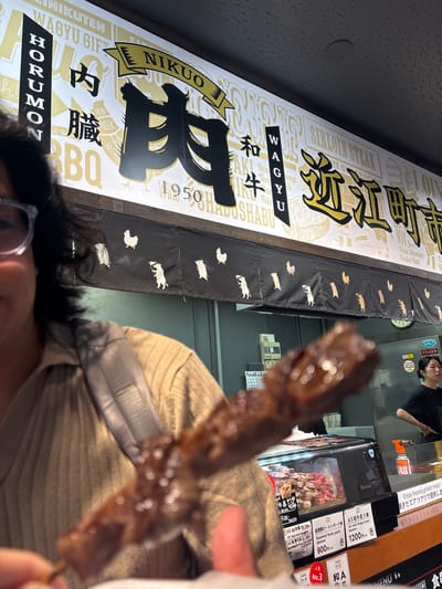 The scene captures an interior view of a bustling food stall, identified as "NIKUO" (肉夫), specializing in meat, located within Omicho Market (近江町市場) in Kanazawa, Japan.
In the foreground, a person's upper body is partially visible on the left, wearing glasses and a light brown, ribbed knit sweater. A gray strap, likely from a bag or camera, crosses their shoulder. Their hand, mostly out of frame, holds a dark, glossy, cooked meat skewer directly in the center, rendered out of focus.
Above and behind the person, a wide illuminated sign prominently displays "NIKUO" in a golden banner. Large black Japanese characters spell "肉" (Niku - meat) and "和牛" (Wagyu - Japanese beef). Additional Japanese text includes "HORUMON" (ホルモン) and "内臓" (Naizou - offal/viscera) on the left, and "近江町" (Omicho) on the right. English text such as "WAGYU GIFT," "SIRLOIN STEAK," and "SHABUSHABU" are also present. The year "1950" is visible, suggesting establishment. Illustrations of various meat cuts like "HEAD," "NECK," "FILLET," and "BREASTS" are interspersed.
Below the main sign, a black fabric curtain, typical of Japanese shop entrances, hangs with a repetitive pattern of white silhouettes depicting chickens and pigs, some pigs holding small flags.
Beneath the curtain, a refrigerated glass display case showcases numerous cuts of raw red meat, likely beef and pork, neatly arranged. Visible price tags include "A5 和牛串焼き" (A5 Wagyu beef skewer) for 1200 yen and "能登豚ローストポーク串" (Noto pork roast skewer) for 900 yen. A red sign indicates "人気 No.3" (Popular No.3).
In the background, behind the counter and display case, a female staff member wearing a black uniform stands with her hands on her hips, looking towards the right. A stainless steel counter surface and various kitchen equipment are visible, including a red spray bottle labeled "GAS."