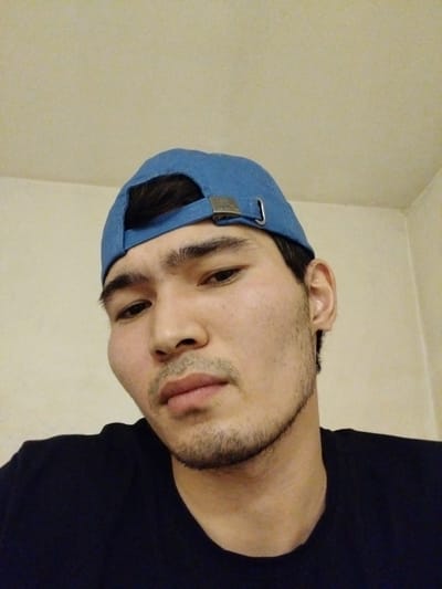 This is a close-up, head-and-shoulders portrait of a young man wearing a blue baseball cap and a black t-shirt. He is looking directly at the camera with a neutral expression. The background is a plain, light-colored wall. The lighting suggests an indoor setting, possibly during the day. The man has dark hair visible under the cap and light stubble on his chin and jawline. His features are prominent, with a broad nose and full lips. There is a metallic buckle on the side of his cap. No specific activity or event is depicted. The setting appears to be a simple room.
