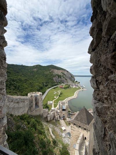 This is a high-angle, medium shot of Golubac Fortress in Golubac Municipality, Serbia, taken on a cloudy day. The foreground is dominated by the rough, gray stone walls of the fortress, creating a natural frame for the scene.

Below the foreground walls, a series of ancient stone structures, including towers with battlements and wooden roofs, are visible. Pathways and staircases wind through the fortress ruins, some with metal railings. Vegetation, including green bushes and grass, grows around the lower levels of the fortress.

Further down, a neatly mown green lawn stretches towards a calm, greenish body of water, likely the Danube River. A paved road runs parallel to the riverbank, bordered by a few small, white buildings. In the distance, a large, tree-covered hill rises steeply, its slopes dotted with more vegetation. The sky above is a mix of blue patches and wispy white clouds. A few distant boats are visible on the water. There are no people clearly visible in the frame.