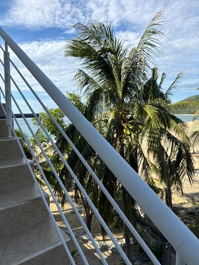 The image captures a view from a tiled staircase with a white metal railing. Beyond the railing, palm trees with green fronds are prominent, with some coconuts visible. The background reveals a sandy beach, the ocean with calm water, and a hazy sky with scattered clouds. In the distance, a green hillside is visible. The scene is likely from a coastal area in Song Cau, Vietnam, given the presence of palm trees and beach scenery. No people are visible in the frame.