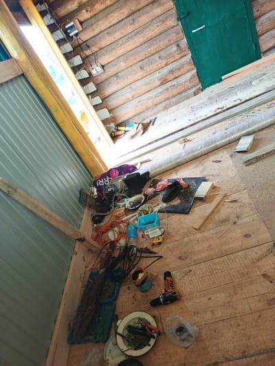 An interior view of a wooden structure, likely a workshop or shed, in Islambaevo, Russia, shows a disarray of tools and construction materials on a wooden plank floor. The room is lit by natural light streaming in from a tall, narrow window frame on the left. A green door is visible on the far wall, constructed of wood planks. In the foreground, a variety of items are scattered: a bundle of twigs resembling a broom, a blue plastic toolbox, various electrical cords, a cordless drill, a white enamel bowl containing tools and fasteners, and a plastic bag. Other objects include a black pair of shoes on a black mat, a roll of material wrapped in plastic, and pieces of lumber. The wooden walls are made of horizontally stacked logs. The floor is covered in sawdust and wood shavings, indicating recent work. No people are present in the image.