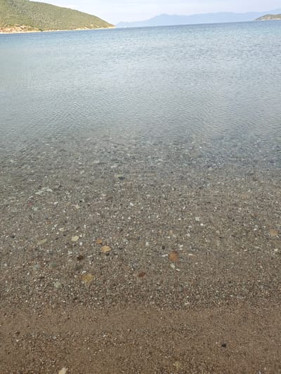 The image captures a static, panoramic view of a clear coastal environment in Nea Peramos, Greece. The foreground features a transition from dry, light brown sand at the bottom of the frame to a wet sandy beach leading into extremely clear, shallow water. Below the water's surface, the seabed is distinctly visible, composed of a mixture of fine sand, various sizes of pebbles, and shell fragments. The water exhibits minor, subtle ripples on its surface, indicating calm conditions.
In the mid-ground, the body of water extends, maintaining its transparency and light blue-grey coloration. The left background is dominated by a verdant, tree-covered hill or low mountain, sloping down towards the water's edge. A lighter-colored, possibly exposed earth or rock band is observable along the base of this landmass. The far background presents distant, hazy blue mountain ranges under a bright, diffused sky, extending across the horizon. No human subjects, man-made objects, actions, or interactions are present within the frame. The scene is entirely natural, focusing on the interface between land and sea.