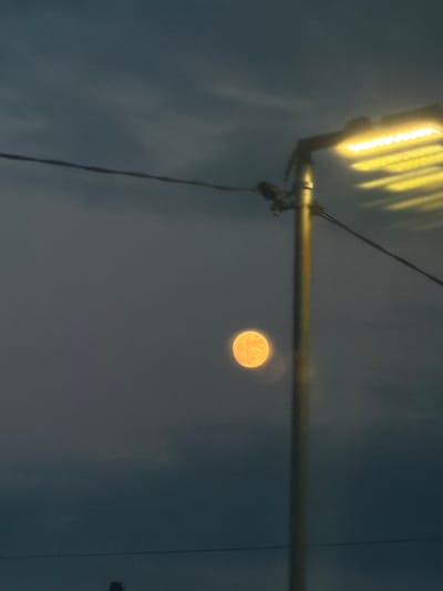 The image captures a nocturnal scene in Michurinsk, Russia, featuring a prominent full moon and an artificial light source set against a dark, hazy sky. The moon, bright and distinctly spherical with visible surface texture, glows in warm yellow and orange tones, positioned on the right side of the frame.
In the upper left quadrant, a luminous artificial light fixture, possibly a street light or a similar elongated light panel, emits a strong, warm yellow glow. This light appears somewhat blurred and streaky, indicating potential motion, reflections, or atmospheric conditions. The fixture is mounted on a dark, horizontal pole that extends across the upper-middle section of the image. Several dark wires or cables hang from this pole, descending into the lower left.
The sky is a muted, dark blue-gray, showing signs of haze or indistinct cloud cover, which contributes to a slightly grainy and soft-focused quality across the image. There are no people visible, nor is any specific activity taking place; the scene is a static composition of natural and urban elements. The time of day is clearly night, with the moon and artificial light providing the primary illumination. No text is visible in the image.