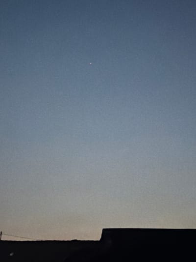 The image depicts a clear sky above a silhouetted rooftop in Denasri Kulon, Indonesia. A single, small, reddish-orange point of light, possibly a star or planet, is visible in the upper center of the frame against the gradient of the twilight sky. The sky transitions from a deep blue at the top to a lighter, warmer hue near the horizon. The foreground is dominated by the dark silhouette of a building's roofline, with a thin horizontal line extending from the left side of the frame, suggesting a utility line. No people or animals are present in the image.