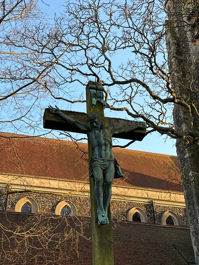 This image depicts a crucifix and church structure in Brentwood, United Kingdom. A central bronze corpus of Jesus Christ, exhibiting a dark green verdigris patina, is mounted on a weathered wooden cross. The figure features a beard, hair, crown of thorns, and a loincloth. A bronze 'INRI' placard is affixed above the figure's head. In the background, a church building with a reddish-brown tiled roof, stone and flint walls, and several arched windows is partially visible. A stone buttress or tower section stands on the right. Numerous bare, dark tree branches are silhouetted against a clear blue sky across the upper and lateral areas. The scene is illuminated by natural daylight.