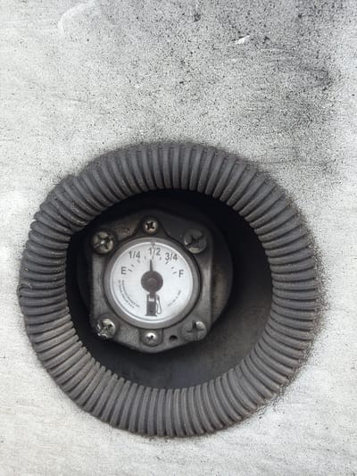 A close-up, overhead view shows a fuel gauge set into a textured, concrete surface. The gauge is circular with a clear glass face, marked with "E", "1/4", "1/2", "3/4", and "F". A thin, black needle points between "1/4" and "1/2", closer to "1/4". The gauge is surrounded by a corrugated black rubber boot, which is set into a circular recess in the concrete. The concrete surface is speckled with dark flecks and some faint, white curved lines. The setting appears to be outdoors, as indicated by the natural lighting and the weathered appearance of the materials. The location is identified as Union, United States of America.