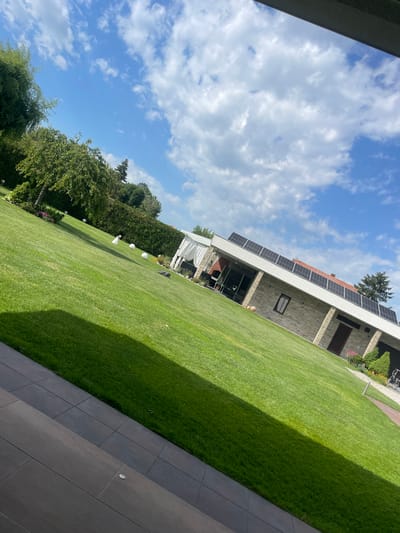 The image shows an exterior view of a building with solar panels on the roof, taken on a sunny day with partly cloudy skies. A well-maintained green lawn stretches from the building towards the foreground, with some landscaping features like trees and decorative objects such as white spherical lights. A patio area with dark-colored paving stones is visible in the foreground. Based on the prompt, this scene is located in Ryahovo, Bulgaria.