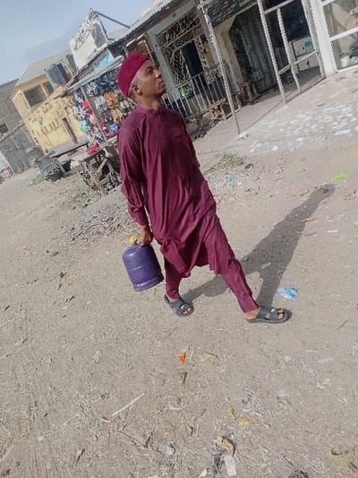 An adult male is observed walking on a dusty, unpaved street in Sharada, Nigeria, on a bright, sunny day. He wears a maroon kaftan, matching trousers, and a maroon skullcap, with dark-colored slide sandals on his feet. He carries a purple, small-to-medium-sized gas cylinder in his right hand, extended downwards. His gaze is directed upwards and to his right. The ground is littered with small debris, including paper, plastic, and organic matter. In the background to the left, market stalls with displayed footwear and other goods are visible, alongside a light yellow building. To the right, storefronts with glass panels and metal grates line the street. Strong shadows are cast on the ground to the man's right, indicating direct sunlight from above and to his left.