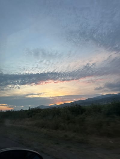 The image displays a captivating sunset or sunrise over a natural landscape in Gavrailovo, Bulgaria, captured from a moving vehicle. The sky dominates the upper two-thirds of the frame, showcasing a striking array of clouds. A prominent horizontal band of clouds across the middle is brightly illuminated by the low-lying sun, glowing in warm tones of gold, orange, and soft pink, contrasting with darker grey and white clouds above and below. Further up, the sky transitions to a lighter, hazy blue-grey with subtle cloud patterns.
In the mid-ground, a series of dark, undulating mountains or hills stretches across the horizon, appearing as silhouettes against the vivid sky, with hints of the golden light catching their peaks. The foreground consists of dark, indistinct grassy or bushy terrain, suggesting a rural or undeveloped area. A small, dark, curved object, likely part of a car door or mirror, is visible in the bottom right corner, indicating the photo was taken from inside a vehicle. The slightly blurred foreground elements further suggest motion. The scene beautifully captures the tranquil and colorful transition of day to night or night to day in a scenic, mountainous region. No visible text is present in the image.