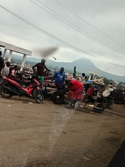 The scene is an outdoor area in Itobe, Nigeria, under an overcast sky. The ground is composed of packed earth and gravel. In the background, green mountains rise beneath the cloudy sky, with light-colored concrete structures and a solar street light pole visible at their base. Multiple power lines traverse the upper portion of the frame.
Approximately ten to fifteen adult males are present. On the left, a red motorcycle with black wheels is parked. Several men are gathered around it, standing and observing. In the center foreground, two men are actively engaged in work related to vehicle tires; one man in a red top and red shorts is bending over a detached tire, while another man in a green shirt and red shorts operates a small, wheeled piece of machinery adjacent to it. A dark plume of smoke rises in the mid-ground behind these working individuals. To the far right, additional individuals are visible near stacks of used tires. The overall environment suggests an informal commercial or service hub.
