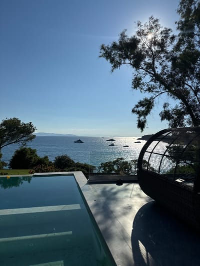 This image depicts a serene coastal view from a modern villa in Vouliagmeni Municipal Unit, Greece. In the foreground, a clean-lined infinity pool with clear, blue water reflects the sky and surrounding trees. The pool's edge is defined by light-colored stone. Adjacent to the pool, a contemporary outdoor lounge area features a large, dark woven pod-shaped chair casting a long shadow on the tiled patio.
The middle ground transitions to a lush green landscape with trees and bushes, leading to the sparkling blue sea. Several yachts and smaller boats are scattered across the water, some appearing as distant silhouettes against the bright, sun-drenched surface. The sun is high in the sky, partially obscured by the branches of a large tree in the upper right quadrant, creating a lens flare effect. In the background, a distant landmass with mountains is visible on the horizon under a clear, bright blue sky. The overall atmosphere is one of luxury, tranquility, and a Mediterranean summer day.