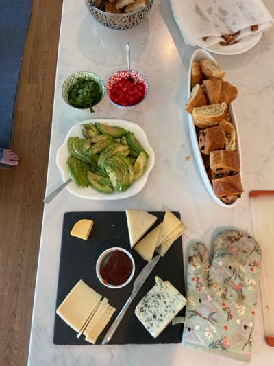 The image shows a kitchen countertop with various foods and objects arranged on it. The countertop is made of white marble, with a light wood floor visible in the background. In the center of the countertop, a large white oval dish filled with sliced avocado is visible. To the right of the dish, a small white oval dish filled with small, golden-brown pastries is positioned. Below the dish, a slate cutting board is placed. The cutting board contains five pieces of yellow cheese, one piece of blue cheese, and a small, red ceramic ramekin filled with red jam. To the left of the avocado dish, a smaller, white ceramic bowl filled with chopped parsley is positioned. Next to the parsley bowl, a red ceramic bowl containing diced red peppers sits on the countertop. Next to the small ceramic bowl, a metal spoon rests on the countertop. There is also an oven mitt on the countertop, which is printed with a floral pattern and features images of koalas. In the upper right corner of the image, a white, rectangular paper towel is sitting on the countertop. The paper towel is partially covered with a plate of bread crumbs. These items suggest that someone is preparing a meal in the kitchen. The location is Rye, New York, United States, based on the provided coordinates.