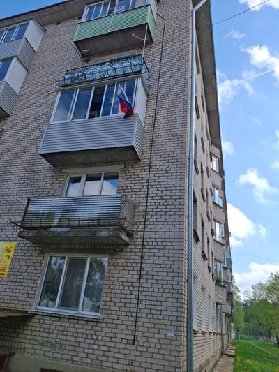The image shows the exterior of a brick apartment building in Ozyorny, Russia. The building is constructed of light-colored bricks. Several windows are visible, some with projecting metal structures that appear to be small enclosed balconies or storage spaces. One window features a small Russian flag. A metal rain gutter runs along the top of the building. The sky is visible above the building. Trees and grass are visible further in the distance.