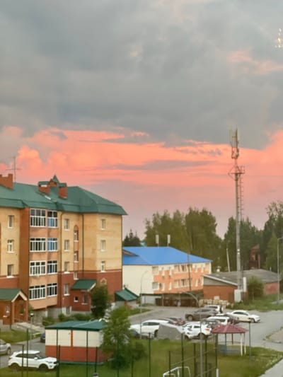 The image shows a residential area in Mezhdurechenskiy, Russia, during a cloudy sunset with pink hues in the sky. In the foreground, a white SUV is parked on a paved surface next to a small, red and white building with a green roof. Beyond this, several other cars are parked on the street, including a dark-colored SUV, a white sedan, and a few other vehicles that are difficult to identify clearly.
A multi-story apartment building with a green roof and yellow and red brick facade is visible on the left. It has numerous windows, many with white frames. To the right of this building, another structure with a bright blue roof and an orange and white facade stands. Behind these buildings, a line of green trees stretches across the middle ground.
A tall communication tower with multiple antennas is prominently featured on the right side of the image, silhouetted against the sky. In the background, a red brick building is partially visible, and the sky is filled with dramatic clouds tinged with pink and gray. The overall scene depicts a typical urban or suburban environment at dusk.