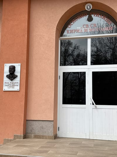 The image displays the entrance of a building, likely a school, located in Chavdar, Bulgaria. The central focus is a white, multi-panel door and window frame set within an arched alcove of a peach-colored stucco wall. The door consists of a white upper section and a lower section with dark window panes.
Above the door, an arched window reflects the dark, bare branches of trees, hinting at a winter or early spring season. Inscribed on the curved glass of this window, in red Cyrillic text, is "ОСНОВНО УЧИЛИЩЕ СВ. СВ. КИРИЛ И МЕТОДИЙ" (Primary School Saints Cyril and Methodius). A small, white loudspeaker is affixed to the wall to the left of the arched window.
Below the entrance, a memorial plaque is mounted on the peach-colored wall. It features a dark, metallic bust relief of a man. The accompanying Cyrillic text identifies him as "Нено Караджов" (Neno Karadzhov), with his lifespan noted as "1928г.-1994г." (1928-1994). The text further states he was a "Учител и директор" (Teacher and Director) "в периода 1954г.-1988г." (during the period 1954-1988). The wall meets a speckled grey concrete section at the bottom right, which then transitions into light brown, rectangular floor tiles. The diffused lighting suggests an overcast day.