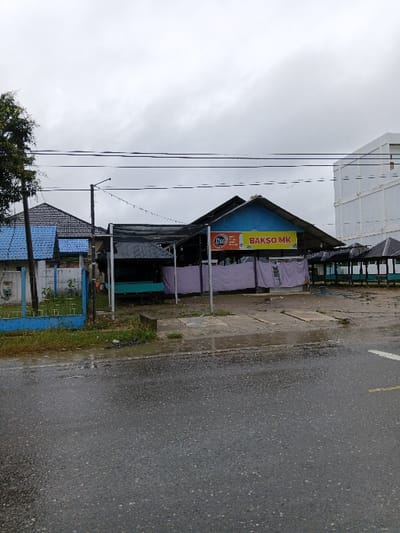 A street scene with a gray sky. A "BAKSO MK" restaurant with a blue and yellow sign. The restaurant's open-air structure has purple curtains or coverings partially drawn. To the left, a white building is visible, obscured by power lines. The street to the right appears wet. A blue and white fence is visible towards the bottom of the frame.