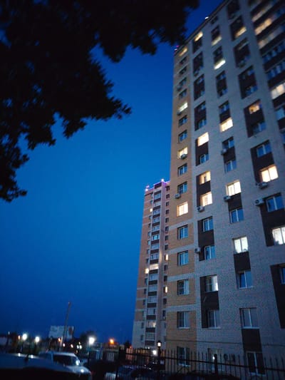 The image shows a nighttime view of two apartment buildings in the city of Krutye Vyselki, Russia. The building on the right is taller and is constructed with light-colored bricks and dark vertical accents. Many of its windows are illuminated, suggesting occupancy. The building on the left is slightly shorter and appears to have a colored light strip along its upper edge. A large, dark tree silhouette is in the foreground on the left, obscuring the top of the sky and the left side of the taller building. The sky is a deep blue, indicative of twilight or night. In the lower portion of the image, a fence is visible, and beyond it, car headlights and streetlights illuminate a dimly lit street or parking area. Some vehicles are partially visible. The scene depicts a residential area at dusk or night.