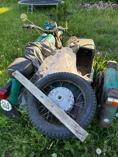 The image depicts a dilapidated green motorcycle with a sidecar abandoned in a grassy area, presumably in Barziya, Bulgaria. The motorcycle is heavily rusted and in disrepair, with a missing seat and several components covered in debris. A wooden plank rests against the sidecar wheel. The environment is overgrown with grass and dandelions, suggesting neglect. There are no people visible in the image.