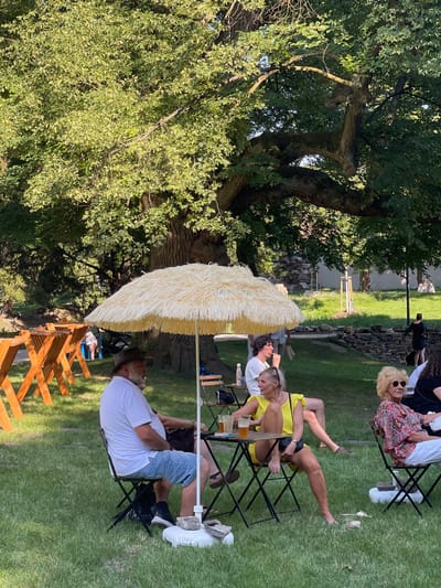 In a park setting in Stupava, Slovakia, bathed in the dappled sunlight filtering through a large, leafy tree, a group of people are gathered outdoors. In the foreground, a man wearing a white polo shirt, denim shorts, and a hat sits in a folding chair under a large, straw-like umbrella. Beside him, a woman in a bright yellow dress is seated, her legs crossed, also under the umbrella. They are seated at a small table with drinks. To their right, another woman with blonde hair and sunglasses wears a colorful patterned shirt and white pants, sitting in a folding chair. In the background, several wooden tables and benches are arranged, and other individuals are visible, some walking or standing. The ground is covered in green grass, and a stone wall runs along the middle ground, with more greenery and trees beyond. The overall atmosphere appears relaxed and social, possibly an outdoor event or gathering.
