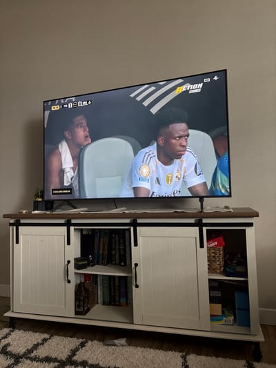 The image displays an indoor residential scene in Tigard, United States, featuring a large flat-screen television placed on a white and brown media console. The walls are light-colored, and a gray and white patterned rug covers the floor in the foreground. The television screen shows a live soccer match with a scoreboard indicating "76:10 | PSG 3 | 0 RMA" and a "COOLING BREAK" notification. The channel logo "ACTION LIVE HD" is visible in the upper right. Two male soccer players are visible on the screen, seated on a bench: the player on the right, identifiable as Vinicius Jr., wears a white Real Madrid jersey with sponsor logos and has a focused expression; the player on the left has a light-colored garment draped over him and an open-mouthed expression. The media console features a dark brown wooden top and white paneled sliding doors with black barn-door style hardware. The left section of the console, partially open, reveals multiple books and a small, ornate gold-colored object. The right section, also open, contains a woven basket filled with various packaged items, and additional objects on a lower shelf. A small potted plant and a small, light-colored circular object are present on the console's top surface to the left of the television. A white sock is visible on the rug in the lower left portion of the frame.